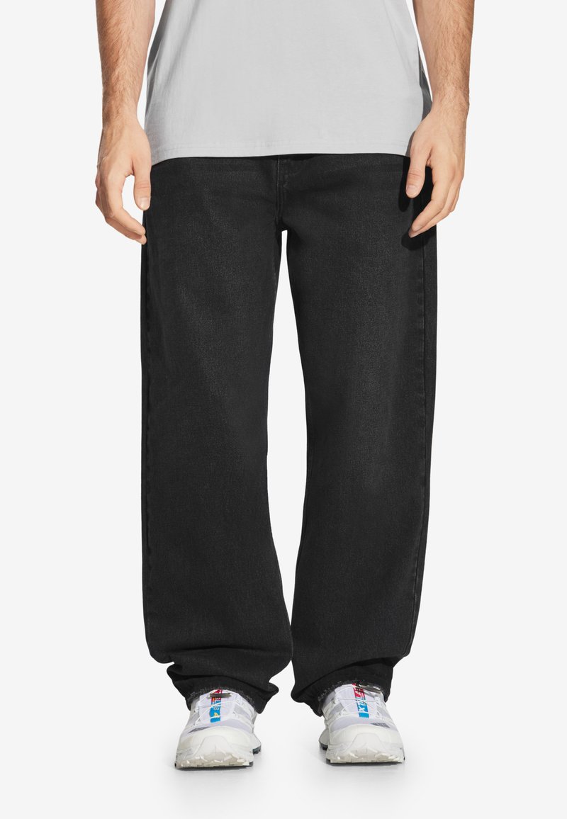 Czarne dżinsy z luźnym krojem, z klasycznym pięcioma kieszeniami. Noszone z białymi butami sportowymi i jasnoszarym t-shirtem.