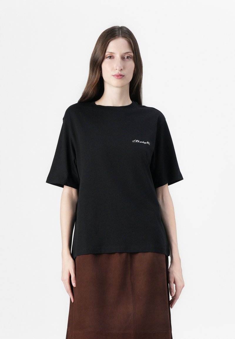 T-shirt di cotone nero con vestibilità ampia, maniche corte e scollo rotondo. Presenta un sottile testo ricamato bianco sull'area sinistra del petto.