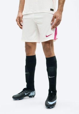 Férfi labdarúgó játékos lábai fehér rövidnadrágban, rózsaszín csíkkal, fekete térdig érő zokniban fehér Nike logóval, és fekete futballcipő kék díszítéssel.