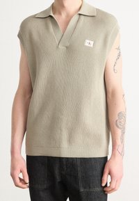 Kurzärmliger, hellbeiger Strickpoloshirt mit V-Ausschnitt, gerippter Textur und einem Logopatch auf der linken Brust. Kombiniert mit dunklen Jeans.