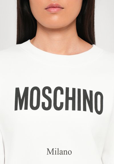 Vit sweatshirt med "MOSCHINO" i feta svarta bokstäver på bröstet, med "Milano" tryckt nedanför i mindre text. Mjuk, slät textur.