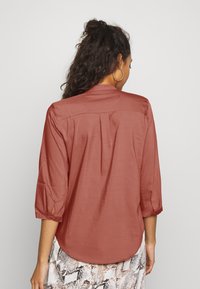 Blouse légère couleur rouille, manches trois-quarts, détail de pli dans le dos et ourlet arrondi. Associée à une jupe texturée à motif.