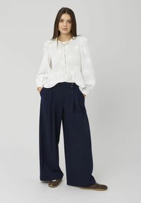 Femme aux longs cheveux bruns portant un chemisier blanc à texture florale et un pantalon large bleu marine, debout les mains dans les poches.