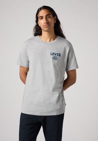 Pilkas medvilninis marškinėlis trumpomis rankovėmis, apvaliu kaklu, su mėlynu Levi's logotipu ant krūtinės ir apvaliu simboliu. Suderinta su tamsiomis džinsomis.