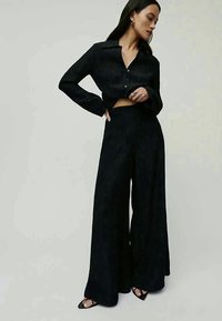 Črna, dolga, zmanjšana srajca s toplim zapenjanjem in reliefno teksturo, v kombinaciji z širokimi hlačami, oba izdelka pa sta okrašena z navpičnimi črtami.