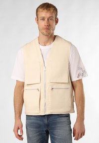 Beige Baumwollweste mit einem Reißverschluss vorne, vier Taschen (zwei Klappen- und zwei Reißverschluss-Taschen) und einer glatten Textur, getragen über einem weißen T-Shirt.