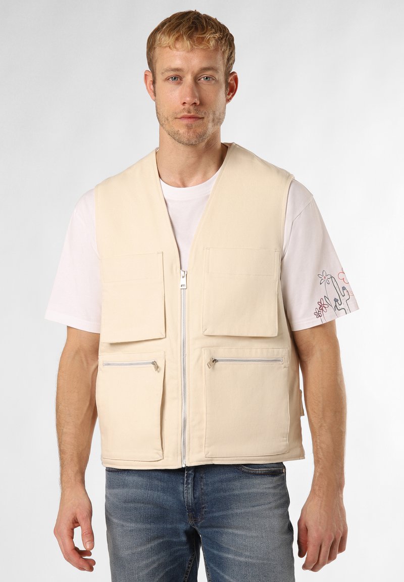 Beige Baumwollweste mit einem Reißverschluss vorne, vier Taschen (zwei Klappen- und zwei Reißverschluss-Taschen) und einer glatten Textur, getragen über einem weißen T-Shirt.