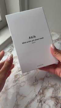 Biały prostokątny box oznaczony "Abib Mild acidic pH sheet mask Aqua fit." Ręce trzymające pudełko na tle marmurowej powierzchni.
