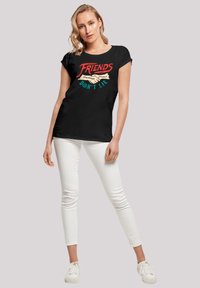 Schwarzes T-Shirt mit roten und türkisfarbenen Texten sowie Designelementen, mit dem Spruch "FREUNDE LÜGEN NICHT", kombiniert mit weißen Slim-Fit-Jeans und Sneakers.