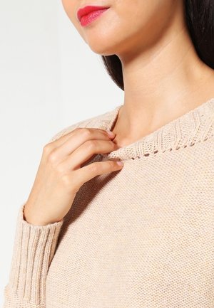 Femme avec un rouge à lèvres rouge ajustant le col d’un pull beige en tricot, gros plan montrant les détails de la main et du cou.
