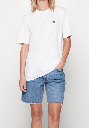 Personne portant un t-shirt blanc uni à manches courtes et un short en denim bleu, une main dans la poche, sur un fond neutre.