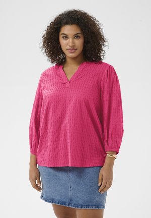 Femme aux cheveux bouclés portant un chemisier rose texturé à manches longues et une jupe en jean bleue, debout devant un fond uni.