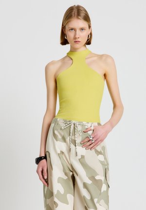 Jeune femme aux cheveux blonds courts portant un top jaune sans manches à col halter et un pantalon cargo camouflage beige avec des anneaux et un bracelet noir.