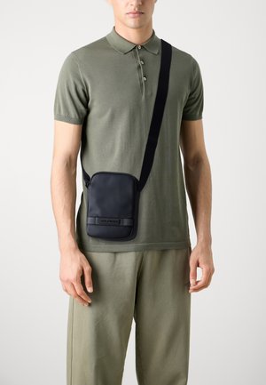 Sort crossbody taske med en glat tekstur, der har en rektangulær form, justerbar skulderstrop og logoaccent. Båret over en olivengrøn polo-shirt.