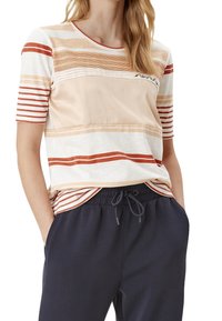 Top de manga corta a rayas en colores crema, beige y óxido. Mezcla de algodón suave con cuello redondo y un sutil detalle de logo. Combinado con joggers oscuros.