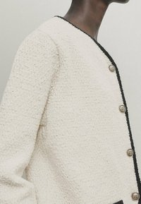 Kurtka tweedowa w kremowym kolorze z czarnymi wykończeniami, z okrągłym dekoltem i pięcioma dekoracyjnymi guzikami z przodu. Szczegóły dotyczące tekstury materiału.