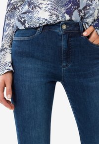 Vaqueros de mezclilla azul oscuro con corte ajustado, que cuentan con un cierre frontal de botón, costuras visibles y una cintura de altura media. Textura suave.