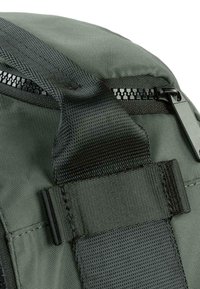 Mochila de tela gris con superficie texturizada, amplia correa negra y cremallera con herrajes negros y un tirador en el lateral.