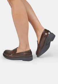 Smeđe kožne cipele na slip-on s niskim složenim petom, s zaobljenim vrhom i dekorativnim šavovima na gornjem dijelu. Teksturirana crna podloga.