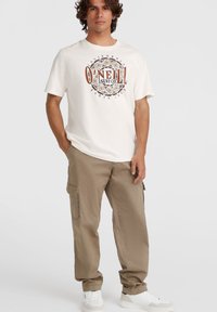 Hvid kortærmet T-shirt med et cirkulært grafisk design, der viser "O'Neill Surf Co" i brun og sort. Kombineret med beige cargo bukser og hvide sneakers.