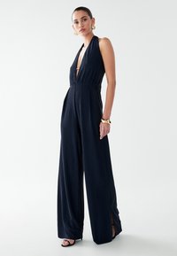 Tamsiai mėlynas halterio stiliaus kombinezonas su giliu V formos iškirpte, plačiomis kojomis, šoninėmis skeltukėmis ir surinktą juosmenį suteikiančia lygia tekstūra. Įskaičiuoti auksinės spalvos aksesuarai.