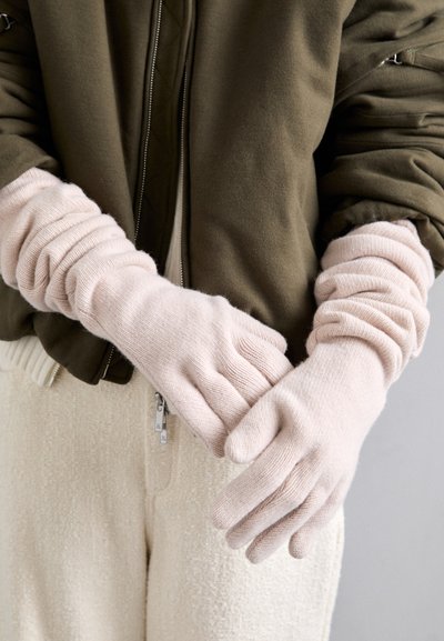 Guantes de lana de color rosa claro con un diseño de canalé, que presentan una textura suave y puños fruncidos, llevados sobre una chaqueta bomber verde oliva.