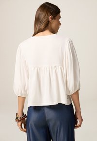 Blusa blanca con mangas abullonadas, detalle fruncido en la parte trasera y adornos de encaje en el dobladillo, combinada con pantalones de mezclilla azul oscuro.