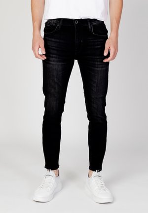 Homme portant un jean slim noir et des baskets blanches, debout devant un fond clair uni.