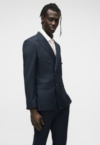 Homme portant un costume croisé bleu marine foncé avec une chemise blanche et une cravate beige clair, debout devant un fond clair uni.