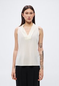 Αμάνικη κρεμ μπλούζα με βαθύ V-λαιμόκοψη και στρωμένο γιακά. Λείο ύφασμα και άνετη εφαρμογή, συνδυασμένη με μαύρο παντελόνι.