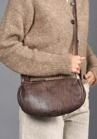 Bolso de cuero marrón cruzado con forma redondeada, superficie texturada y sutil detalle de costura, sostenido por una persona que lleva un cárdigan beige.