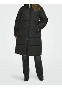 Persoon gekleed in een lange zwarte pufferjas met voorrits, handen in de zakken, gecombineerd met zwarte spijkerbroek en zwarte schoenen, staande op een witte achtergrond.