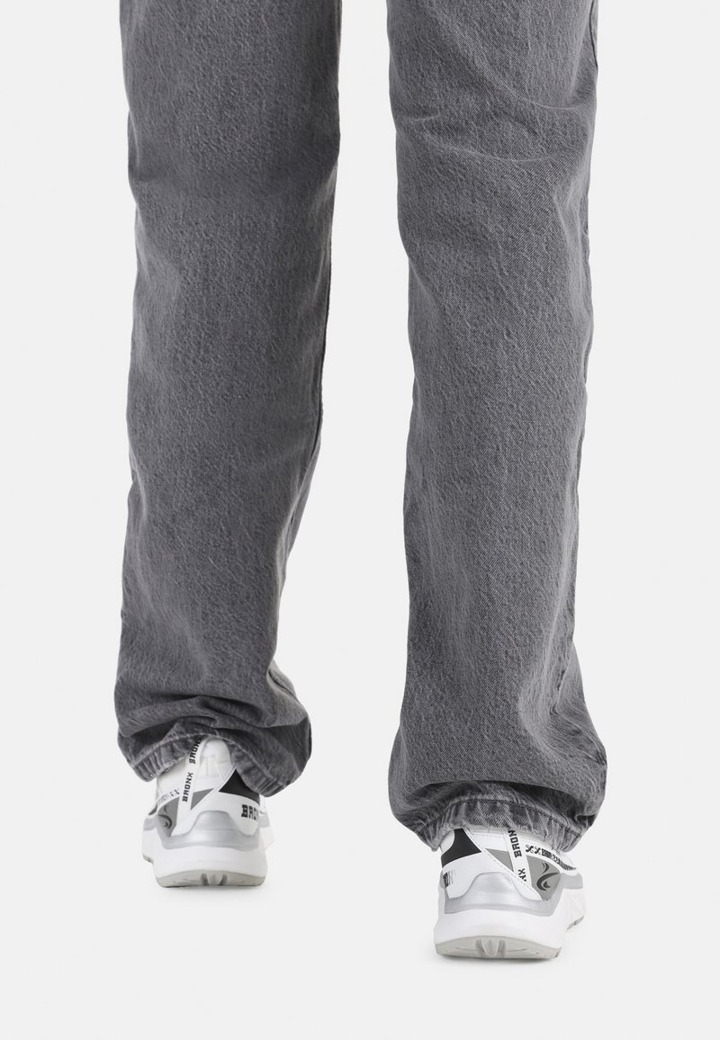 Persona con pantalones grises sueltos y zapatillas blancas de pie sobre un fondo blanco, vista desde atrás a nivel de pantorrilla.