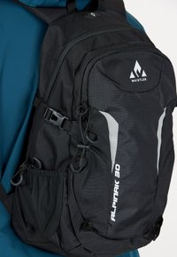 Svart ryggsäck tillverkad av slitstarkt material med en strukturerad yta. Har justerbara remmar, ett meshnät sidofack och reflekterande detaljer.