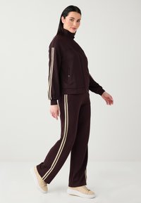 Ensemble de survêtement de sport bordeaux avec des bandes latérales dorées, comprenant une veste zippée, des poches latérales et un pantalon à coupe décontractée associé à des baskets beige.