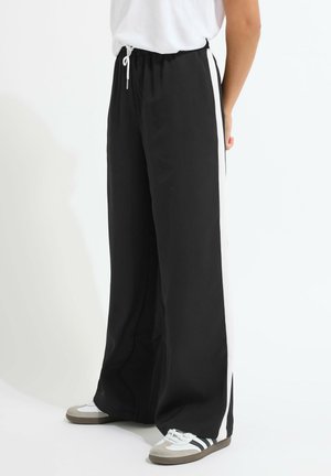 Person iført sorte bukser med vide ben og hvide sidelommer, hvid snøring i taljen, hvid T-shirt og hvide sneakers med sorte striber.