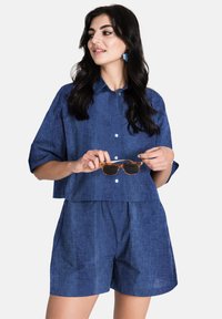 Blå linneskjorta med kort ärm och knäppning fram, parat med matchande blå shorts. Personen håller i solglasögon.