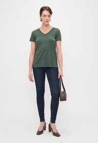 Grøn V-hals T-shirt lavet af blødt stof, parret med mørke skinny jeans og grå hæle. Holder en lille brun håndtaske. Enkel og tætsiddende design.