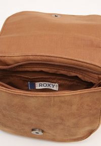 Brązowa torba z materiału o gładkiej fakturze, z zamknięciem na suwak i zapięciem na nap. Wnętrze zawiera metkę z napisem "ROXY" w białym i niebieskim kolorze.