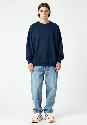 Donkerblauwe oversized sweatshirt met geribde manchetten en zoom, gecombineerd met lichtblauwe baggy jeans en witte sneakers met rode en blauwe accenten.