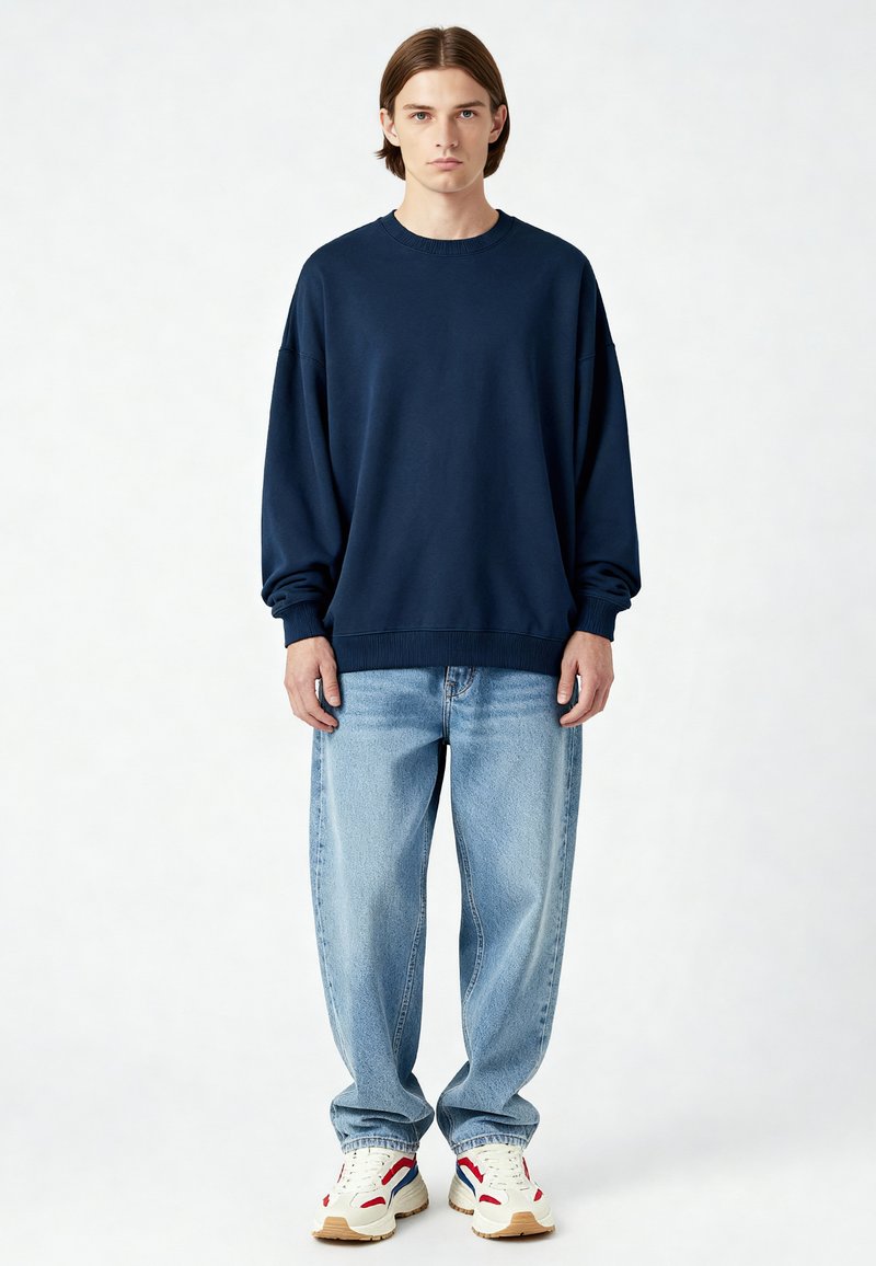 Tamsiai mėlynas per didelis džemperis su raštuotais manžetais ir apatine dalimi, derinamas su šviesiai mėlynomis plačiomis džinsomis ir baltomis sportinėmis bateliais su raudonais ir mėlynais akcentais.