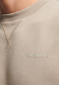 Felpa beige con tessuto testurizzato, dotata di scollatura rotonda e cuciture tono su tono, con il logo "SUPERDRY" di colore chiaro sul fronte.
