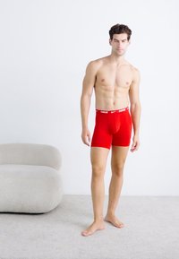 Rode boxershorts van stretchstof met een wit Nike-logo op de tailleband, nauwsluitend ontwerp en lengte tot halverwege de bovenbenen, gedragen door een staande model.