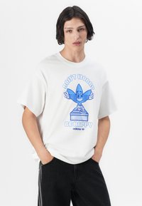 Balta kokvilnas T-krekls ar zilu grafisku attēlu, kurā attēlots smaidīgs tēls ar pārspīlētu Adidas logotipu un tekstu "NEMOKIES, ESI LAIMĪGS".