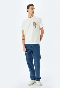 Camiseta de algodón blanca con una flor naranja bordada y hojas verdes en el lado izquierdo del pecho, combinada con jeans de mezclilla azules y zapatillas blancas.