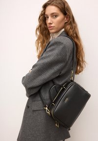 Czarna skórzana torebka z złotymi akcentami, prostokątny kształt, podwójne uchwyty i regulowany pasek, noszona na szarej marynarce w paski.