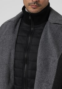 Uomo che indossa un cappotto grigio a spina di pesce sopra un giubbotto trapuntato nero con zip frontale e un alto colletto nero.