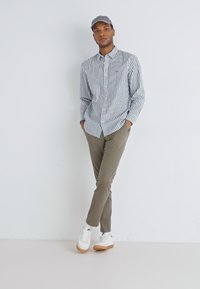 Camicia a righe blu navy e bianca, pantaloni verde oliva e sneakers bianche con suola color caramello, completati da un cappellino grigio.