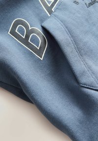 Modrá bavlněná mikina s texturovaným povrchem, obsahující výrazné černé písmeno "B" a další šicí detaily v oblasti kapsy.