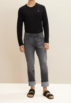 Homme portant une chemise noire à manches longues, un jean gris retroussé et des sandales noires à brides, debout sur un fond neutre.